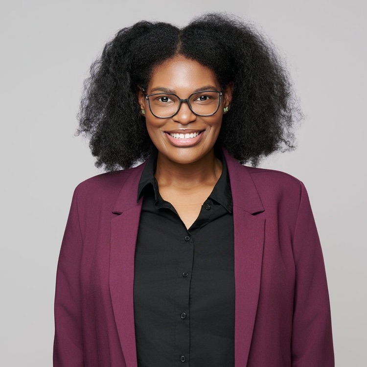 Headshot of a woman in glasses and a plum blazer over a black shirt, light grey background