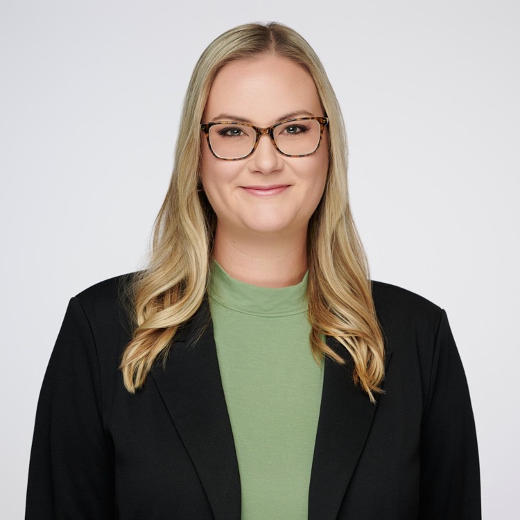 Headshot of a woman in glasses and a black blazer over a sage green top, light background