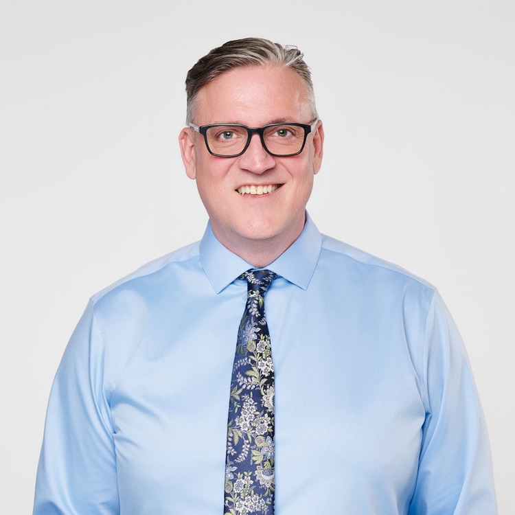 Headshot of a man in glasses, a light blue shirt and patterned tie with no jacket, light background