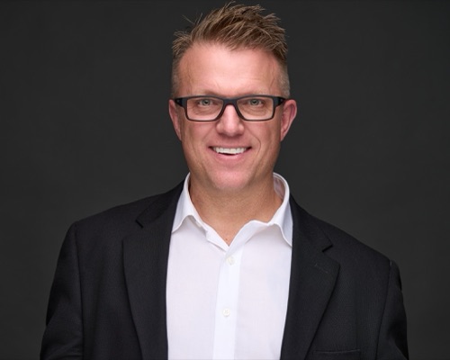 Headshot of a smiling man in glasses and a dark blazer against a dark backdrop, ATX Headshots