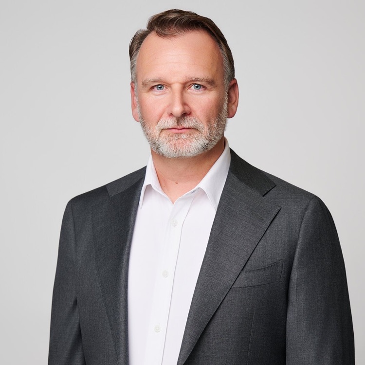 Headshot of a man in a charcoal suit with an open white collar, grey background