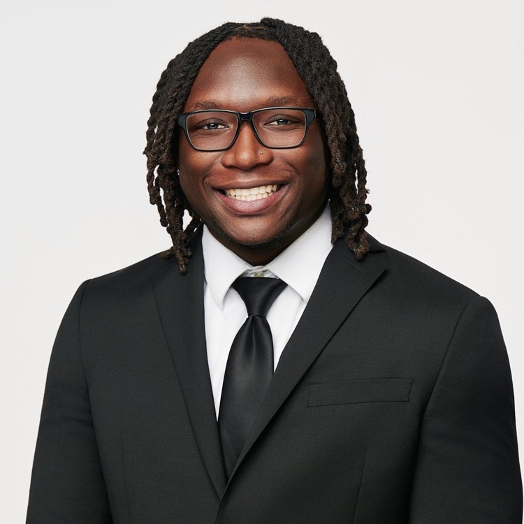 Headshot of a man in glasses, a black suit, white shirt and black tie, light background